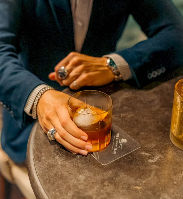 A person's hand, adorned with rings and bracelets, holds an old-fashioned cocktail on a dark table. The coaster reads: THE TENNESSEAN LUXURY HOTEL.