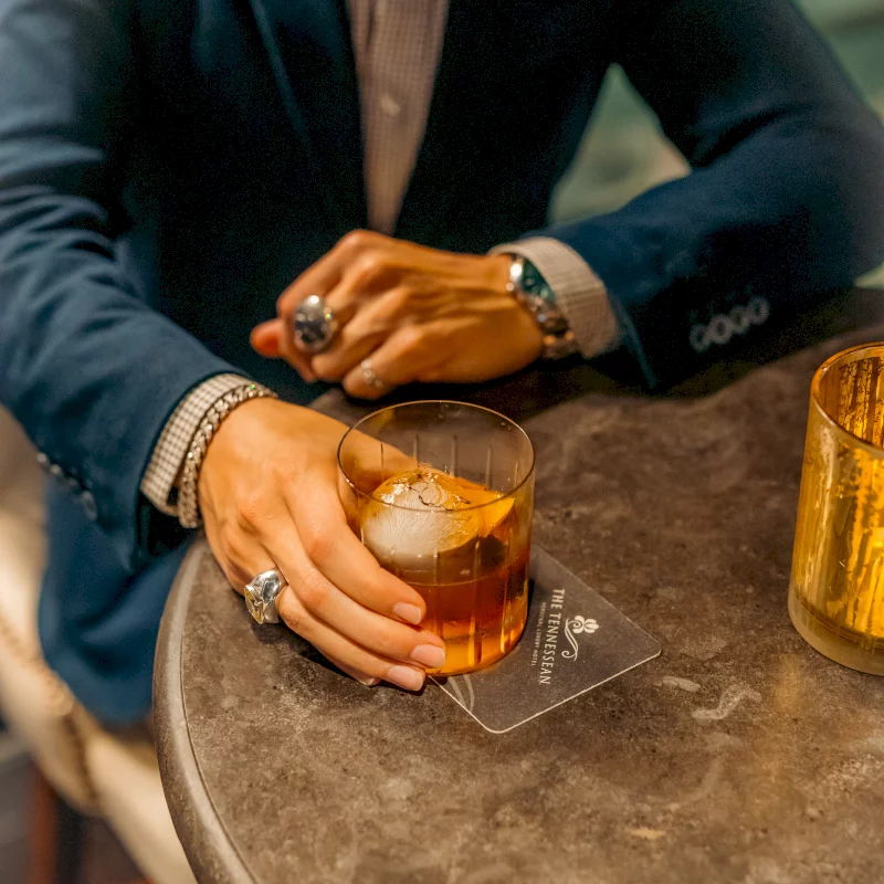 A person's hand, adorned with rings and bracelets, holds an old-fashioned cocktail on a dark table. The coaster reads: THE TENNESSEAN LUXURY HOTEL.