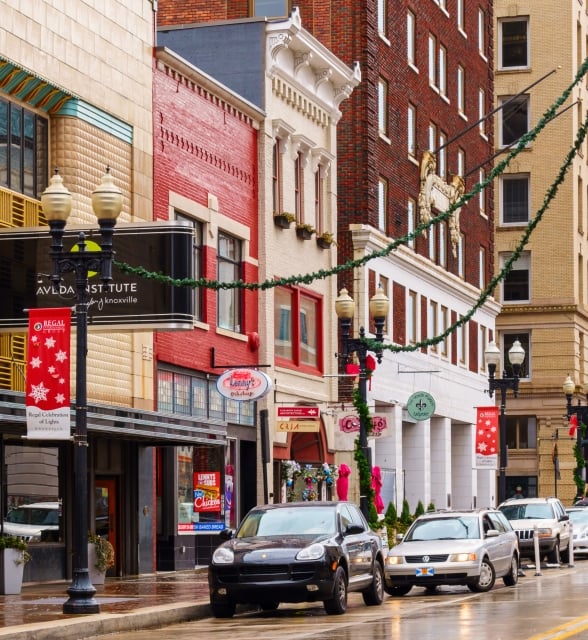 Historic multi-story buildings line a wet downtown street, decorated with garlands and banners. Cars are parked and driving.