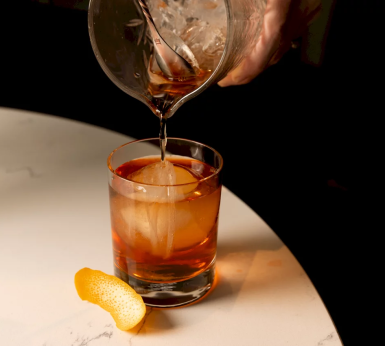 Amber liquid is poured from a mixing glass into a short cocktail glass containing a large ice sphere and an orange peel, resting on a white marble surface.