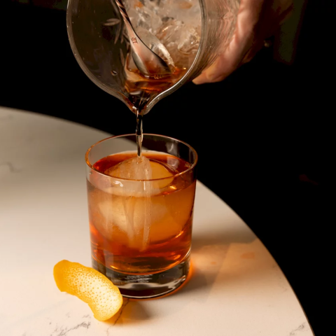 Amber liquid is poured from a mixing glass into a short cocktail glass containing a large ice sphere and an orange peel, resting on a white marble surface.