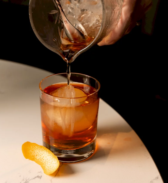 Amber liquid is poured from a mixing glass into a short cocktail glass containing a large ice sphere and an orange peel, resting on a white marble surface.