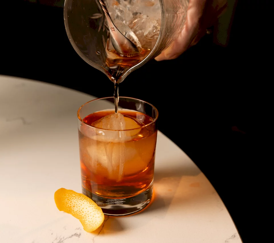 Amber liquid is poured from a mixing glass into a short cocktail glass containing a large ice sphere and an orange peel, resting on a white marble surface.