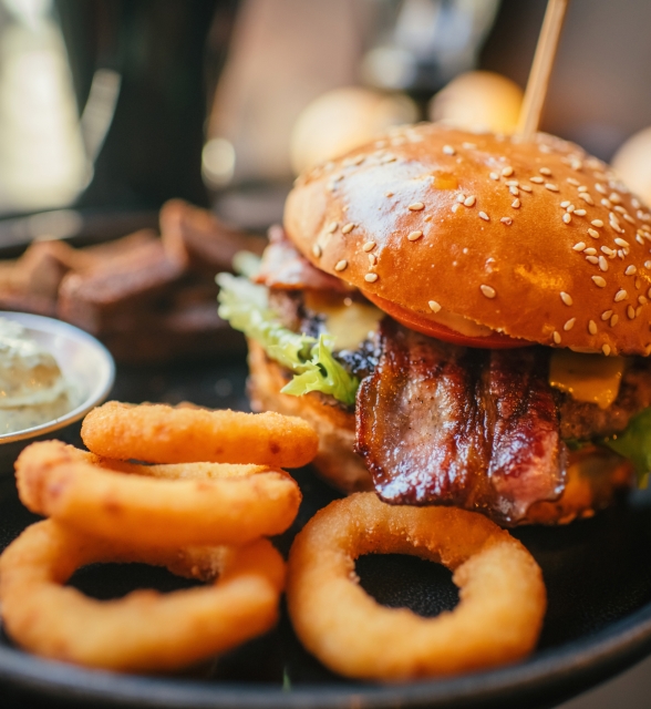 A large, sesame-seed bun burger with bacon, cheese, and lettuce, skewered and presented on a black plate. It is accompanied by golden onion rings, dark bread, and a small bowl of white sauce.