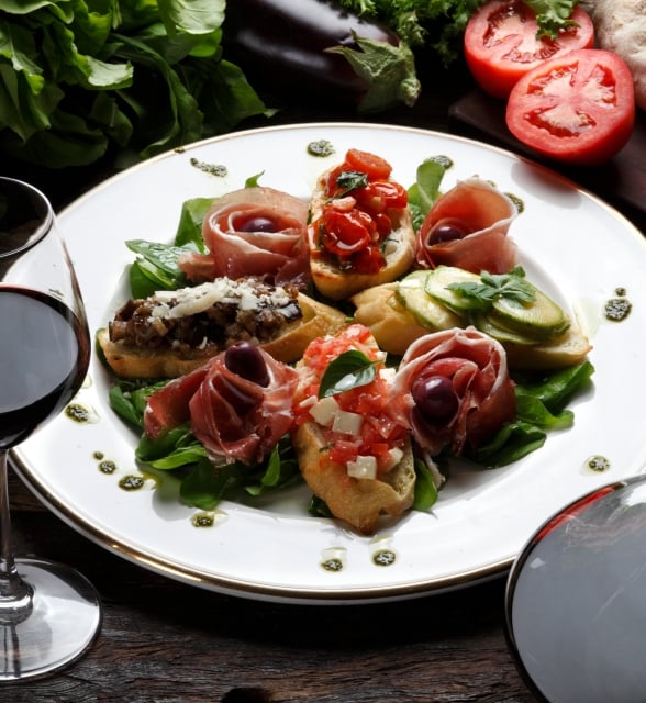 Assorted bruschetta on a white plate with greens and pesto are presented alongside a glass and decanter of red wine. Fresh tomatoes, eggplant, and bread are on a wooden table.