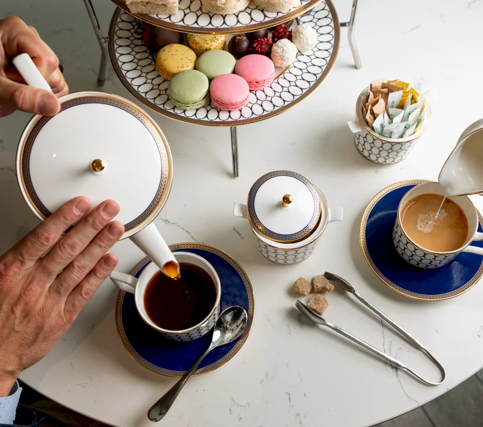 Hands pour tea from a white teapot into a blue cup. Another cup receives milk, next to a tiered stand with colorful pastries on a white marble table.
