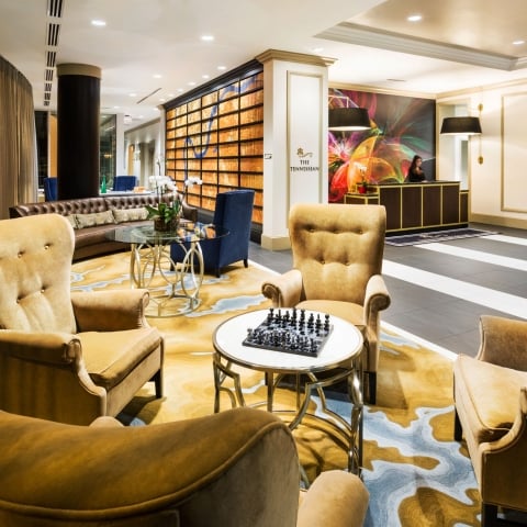 Mustard armchairs surround a chess table on a vibrant patterned rug in an elegant hotel lounge. A reception desk with a clerk stands under a colorful mural. Text: THE TENNESSEAN.