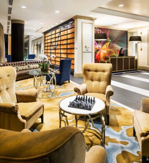 Mustard armchairs surround a chess table on a vibrant patterned rug in an elegant hotel lounge. A reception desk with a clerk stands under a colorful mural. Text: THE TENNESSEAN.