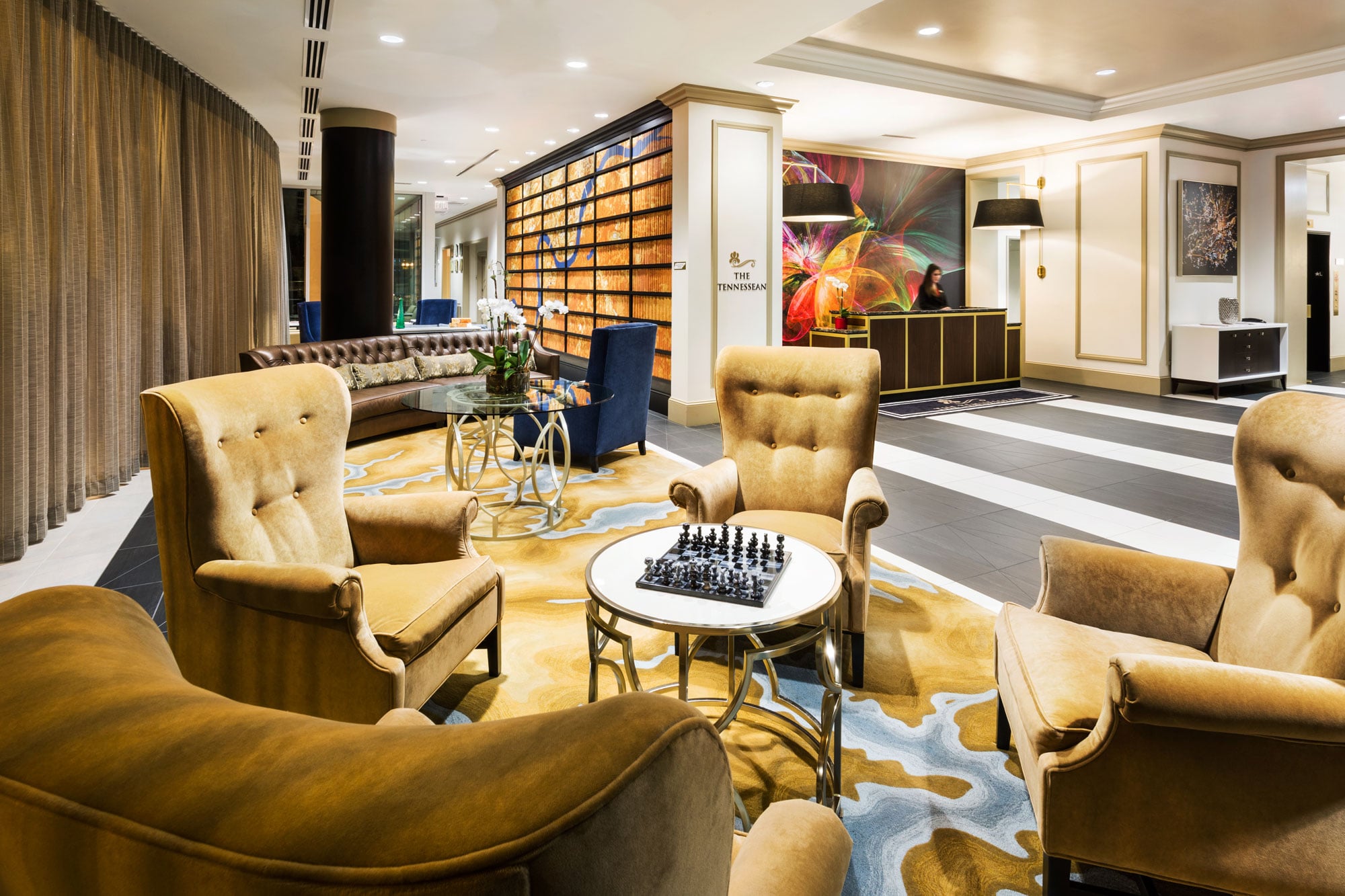 Mustard armchairs surround a chess table on a vibrant patterned rug in an elegant hotel lounge. A reception desk with a clerk stands under a colorful mural. Text: THE TENNESSEAN.