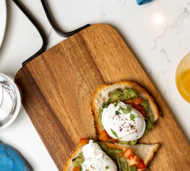Two poached eggs on avocado toast rest on a wooden serving board, set on a white marble countertop with a glass of orange juice.