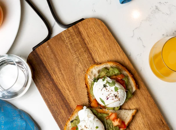 Two poached eggs on avocado toast rest on a wooden serving board, set on a white marble countertop with a glass of orange juice.