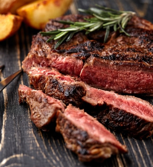 A grilled steak, garnished with rosemary, is partially sliced on a dark wooden cutting board, with potato wedges, a red sauce cup, and a meat fork nearby.