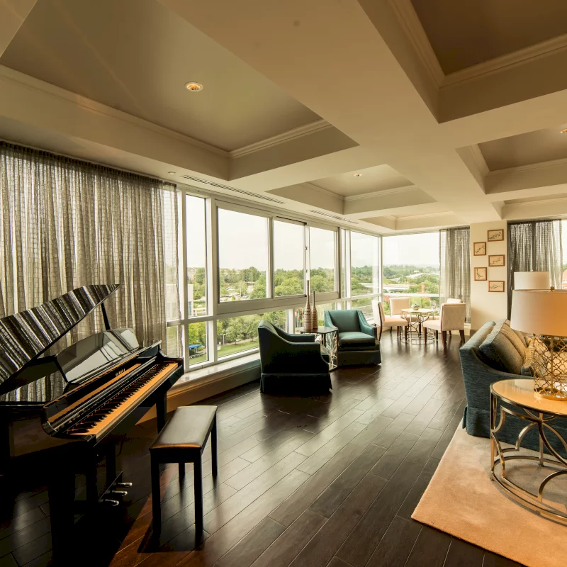 A glossy black grand piano and bench await playing in a sunlit, elegant living room. Dark hardwood floors lead to large windows revealing a lush green landscape and modern seating areas.