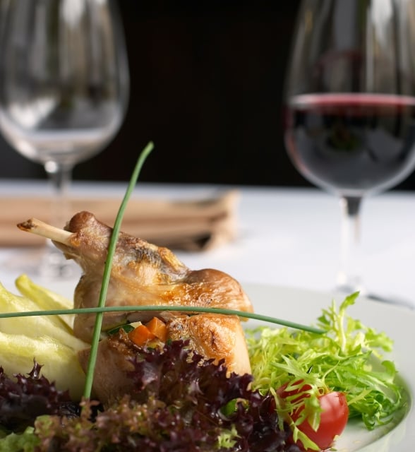 Roasted poultry leg with exposed bone, plated with mixed greens, chives, and vegetables. Two blurred wine glasses, one with red wine, sit on a white tablecloth.