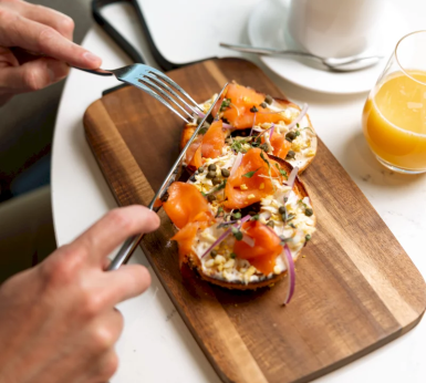 A person cuts a bagel topped with smoked salmon, cream cheese, capers, and red onion, served on a wooden board with orange juice and coffee on a white table.