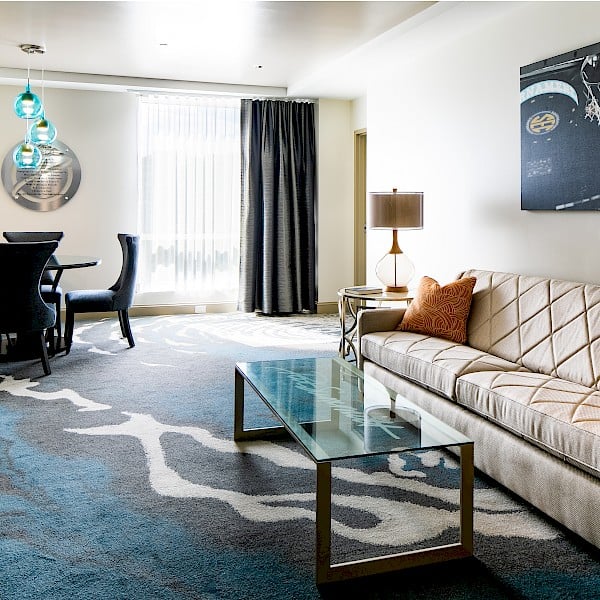 A modern hotel suite is furnished with a beige quilted sofa, glass coffee table, and dining set, overlooking a large window on a blue-grey patterned carpet.