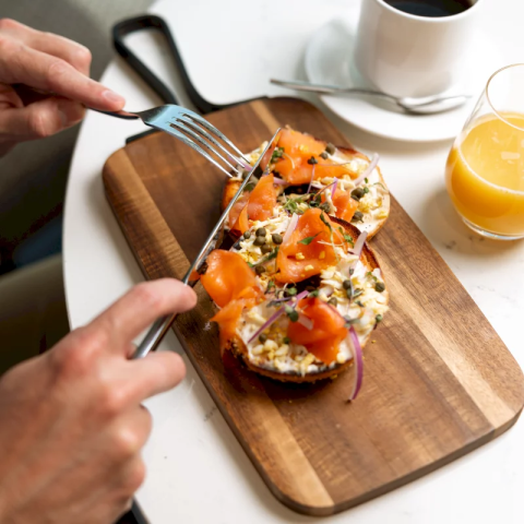 A smoked salmon bagel with cream cheese and capers on a wooden board. Hands cut it with a knife and fork. Coffee and orange juice are nearby on a white table.