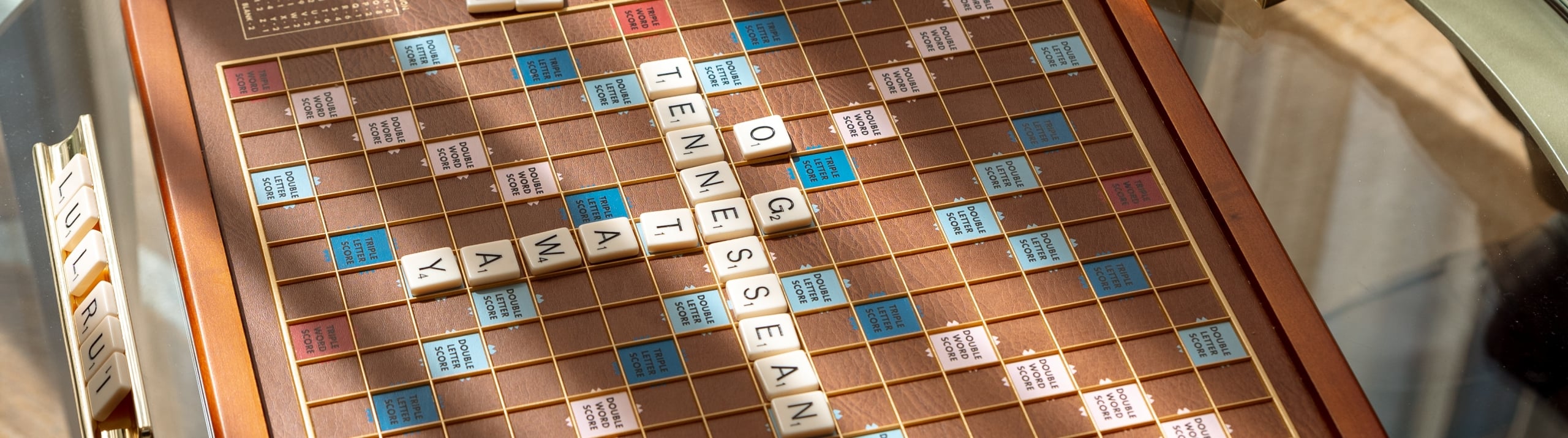 A close-up shot of a classic wooden Scrabble board game on a glass table, spelling out "TENNESSEAN".