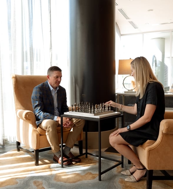 A man and a woman playing a game of chess while seated in large, comfortable armchairs inside a brightly lit hotel lounge.