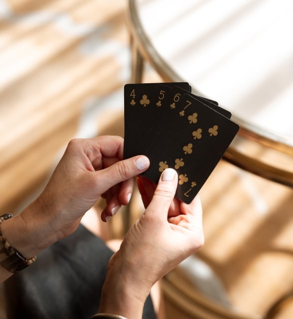 A close-up shot of hands holding five black playing cards with gold lettering at a reflective wooden table.