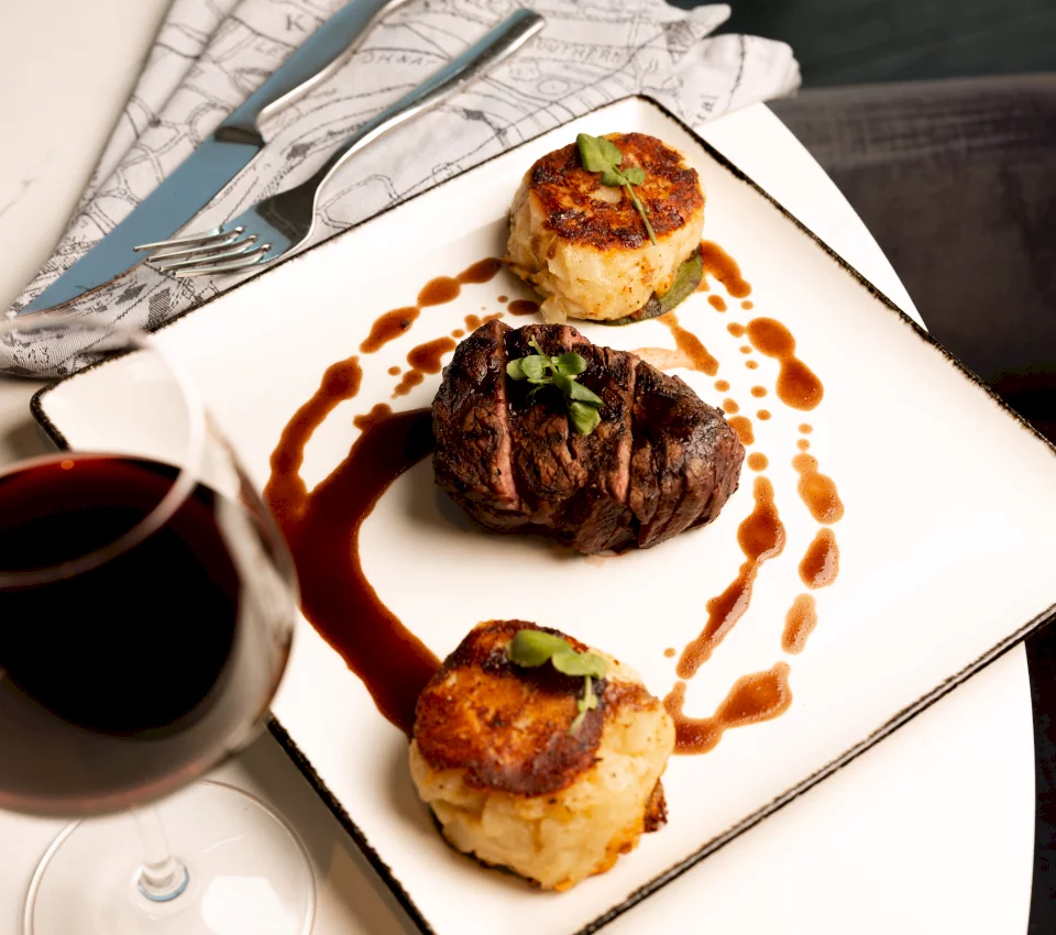 Seared steak and two potato cakes, garnished with microgreens and brown sauce, on a white plate. A red wine glass and cutlery are beside a map-patterned napkin.