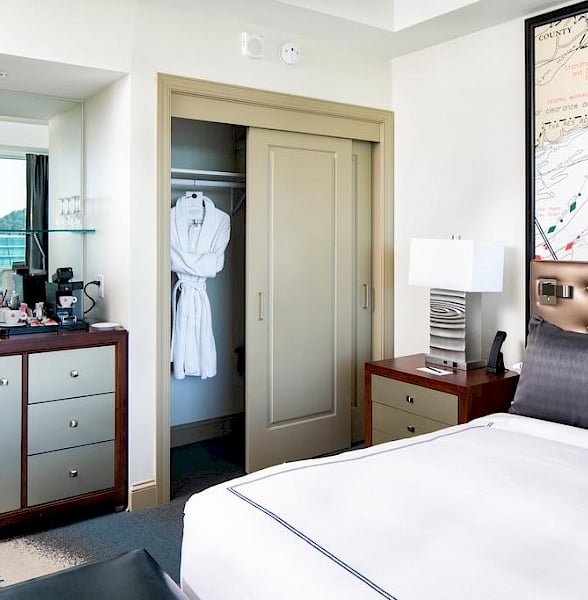 A white bathrobe hangs in a partially open closet of a hotel room, which includes a bed, nightstand with lamp, dresser, and a map with "COUNTY".