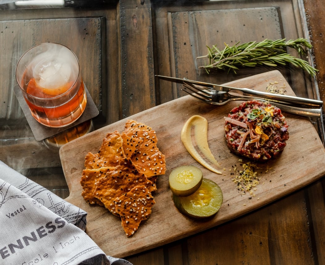 Steak tartare, crackers, and pickles are presented on a wooden board next to a cocktail with mist. This is arranged on a dark table with a map-printed towel showing "TENNESS".