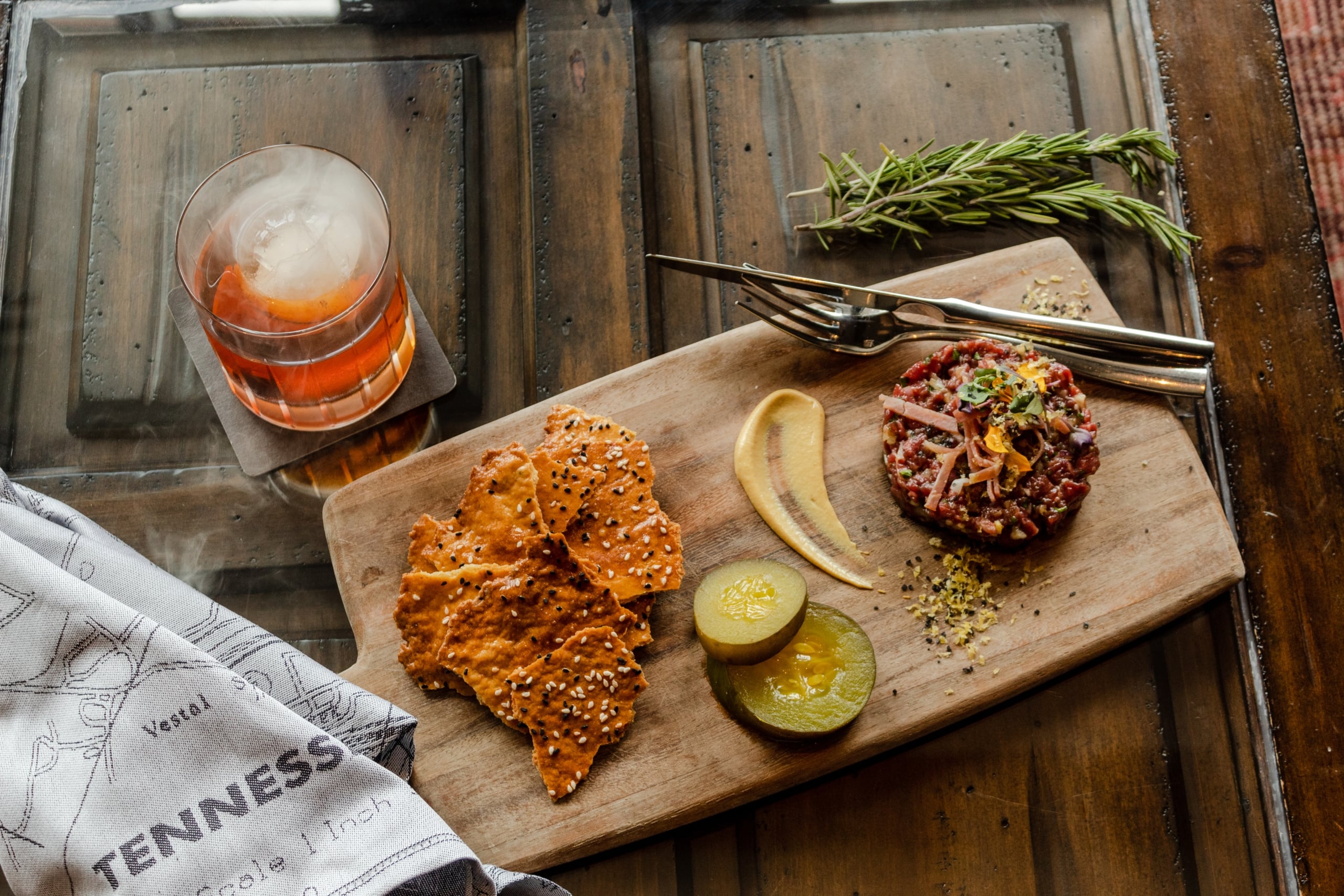 Steak tartare, crackers, and pickles are presented on a wooden board next to a cocktail with mist. This is arranged on a dark table with a map-printed towel showing "TENNESS".