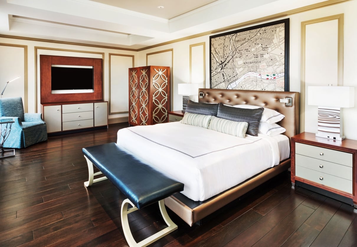 A large bed with white linens and a dark bench at its foot sits in a luxurious bedroom with a framed "KNOXVILLE" map above the headboard and a TV.