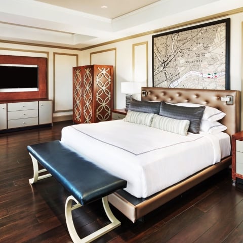 A large bed with white linens and a dark bench at its foot sits in a luxurious bedroom with a framed "KNOXVILLE" map above the headboard and a TV.