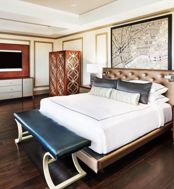 A large bed with white linens and a dark bench at its foot sits in a luxurious bedroom with a framed "KNOXVILLE" map above the headboard and a TV.