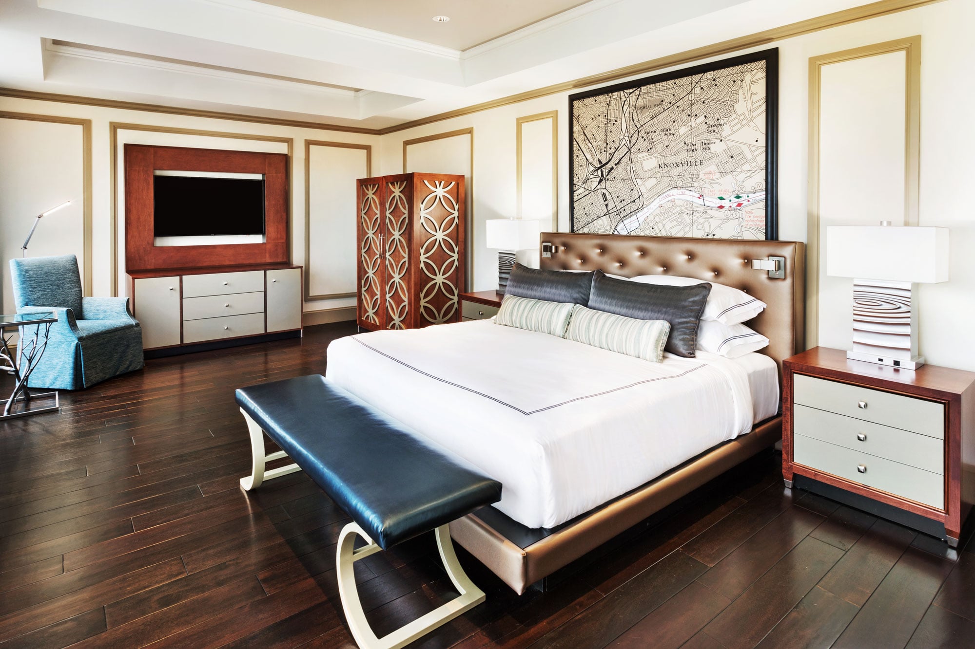 A large bed with white linens and a dark bench at its foot sits in a luxurious bedroom with a framed "KNOXVILLE" map above the headboard and a TV.