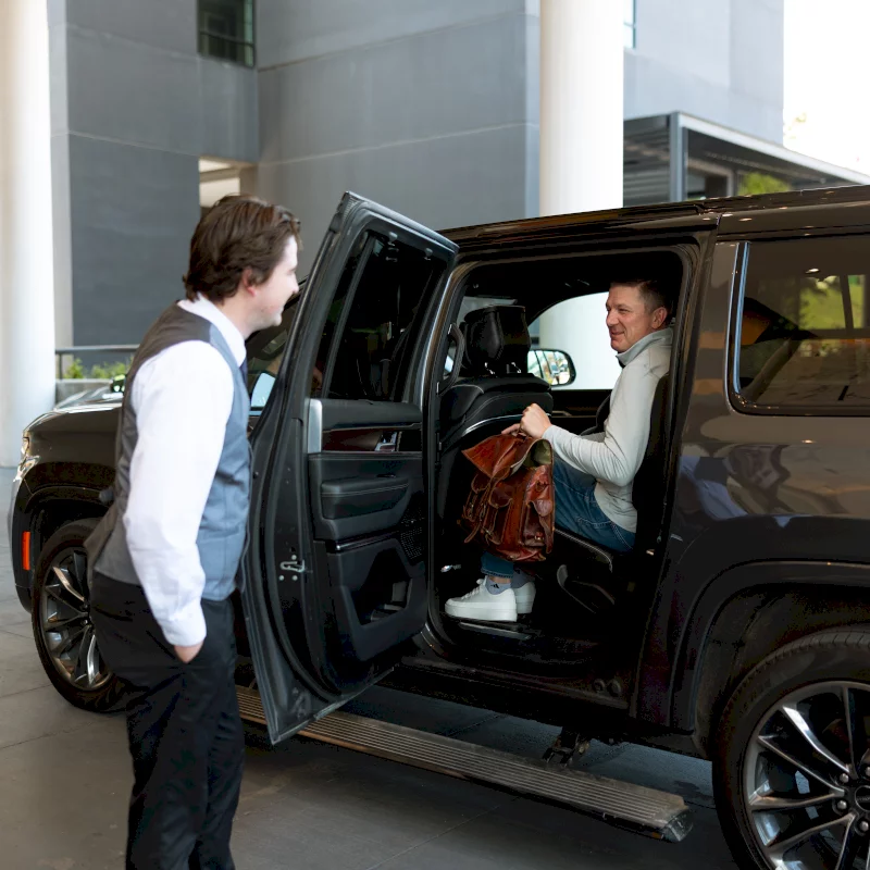 A passenger sits inside a black SUV, holding a brown bag, smiling at a uniformed valet standing by the open door. They are at the entrance of a modern building.