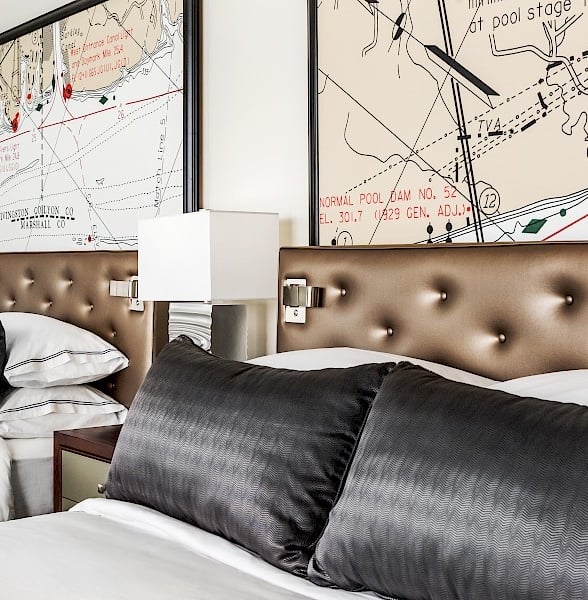 Two beds with dark pillows and brown tufted headboards are arranged under large framed maps.