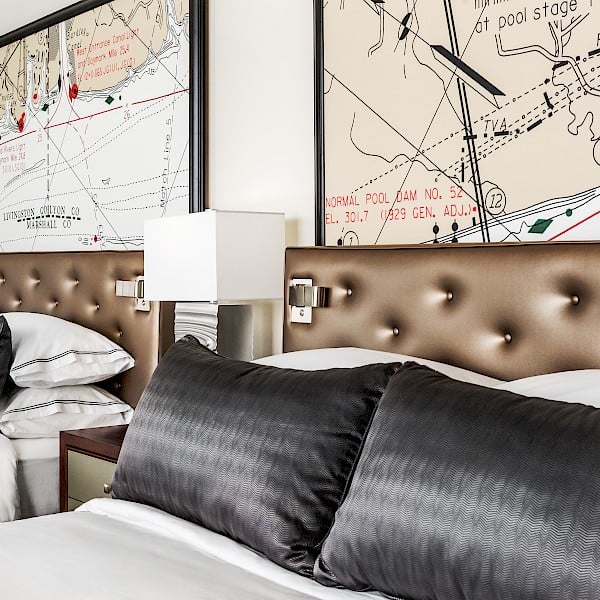 Two beds with dark pillows and brown tufted headboards are arranged under large framed maps.