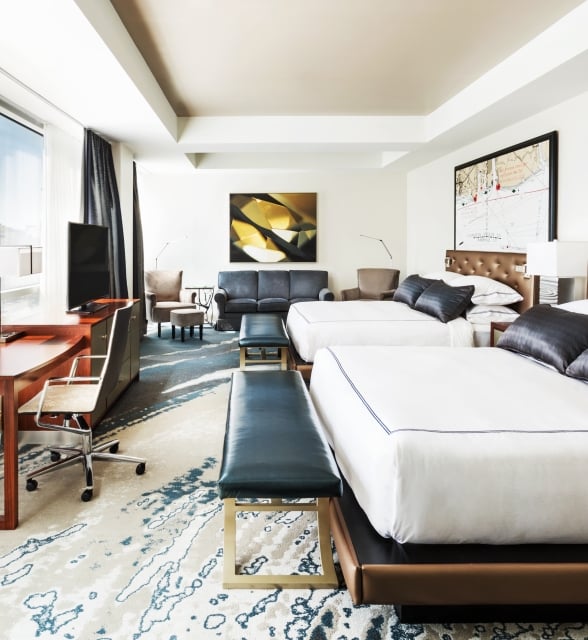 Two beds and a desk furnish a modern hotel room with large windows overlooking a city. Framed maps above the beds include text: "Seven Aerial Power Cross," "NORMAL POOL DAM NO. 52."