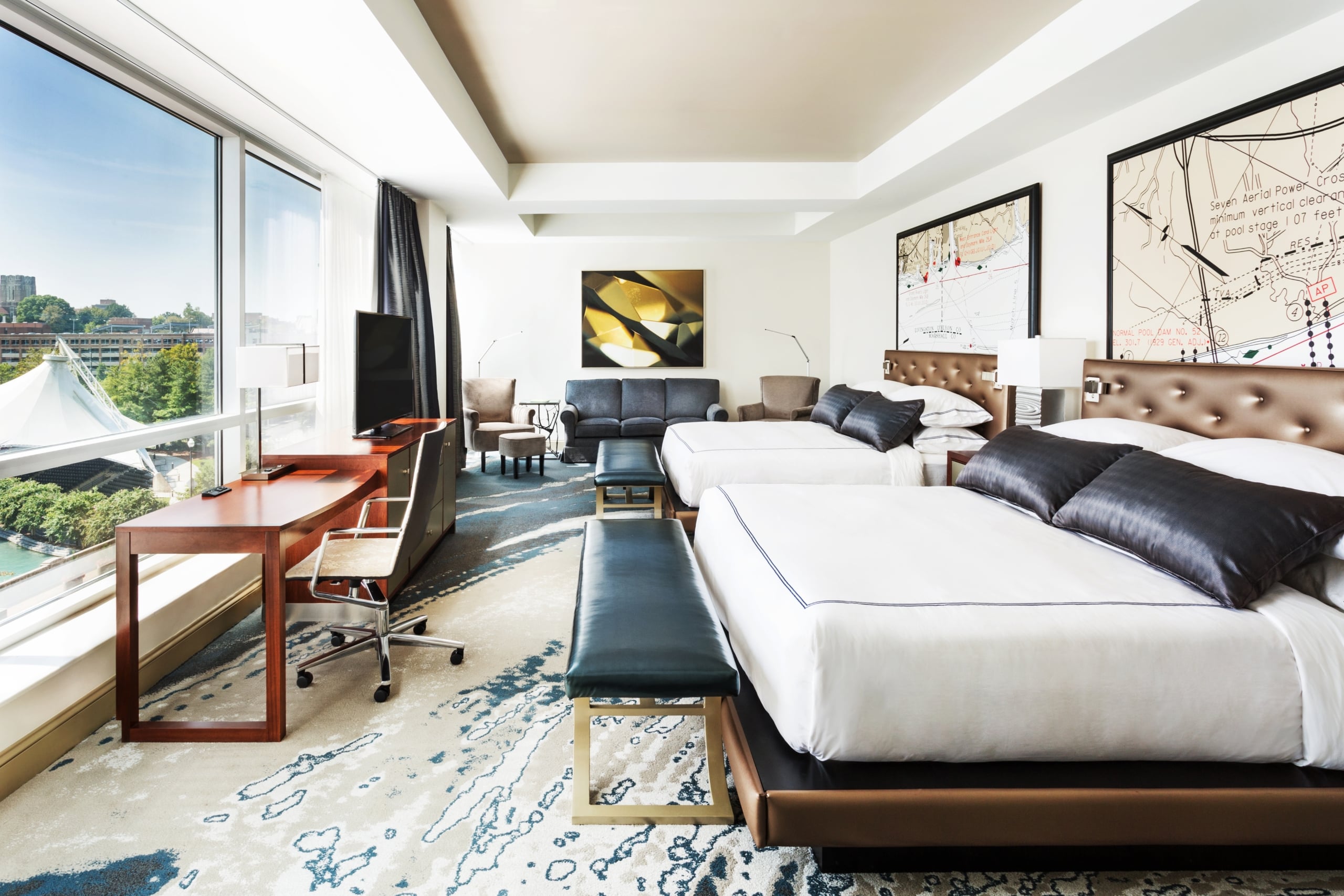 Two beds and a desk furnish a modern hotel room with large windows overlooking a city. Framed maps above the beds include text: "Seven Aerial Power Cross," "NORMAL POOL DAM NO. 52."