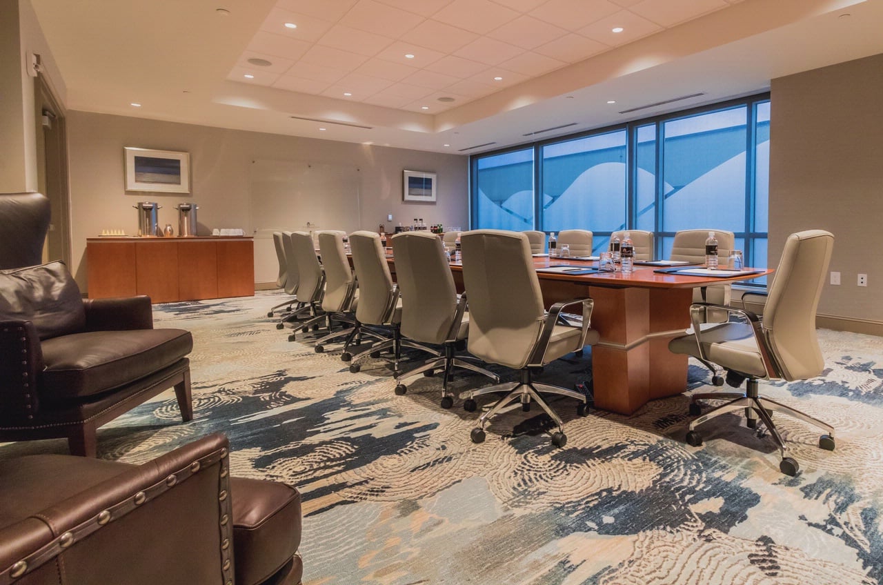 A long wooden conference table with chairs sits ready for a meeting, featuring bottles of ACQUA PANNA and SAN PELLEGRINO, in a modern room with large windows.