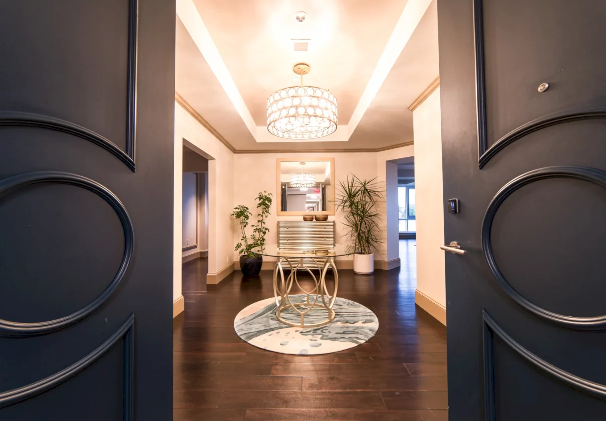 A round glass-top table and ornate chandelier are centered in a grand, brightly lit entryway, viewed through open dark doors, featuring dark wood floors and potted plants.
