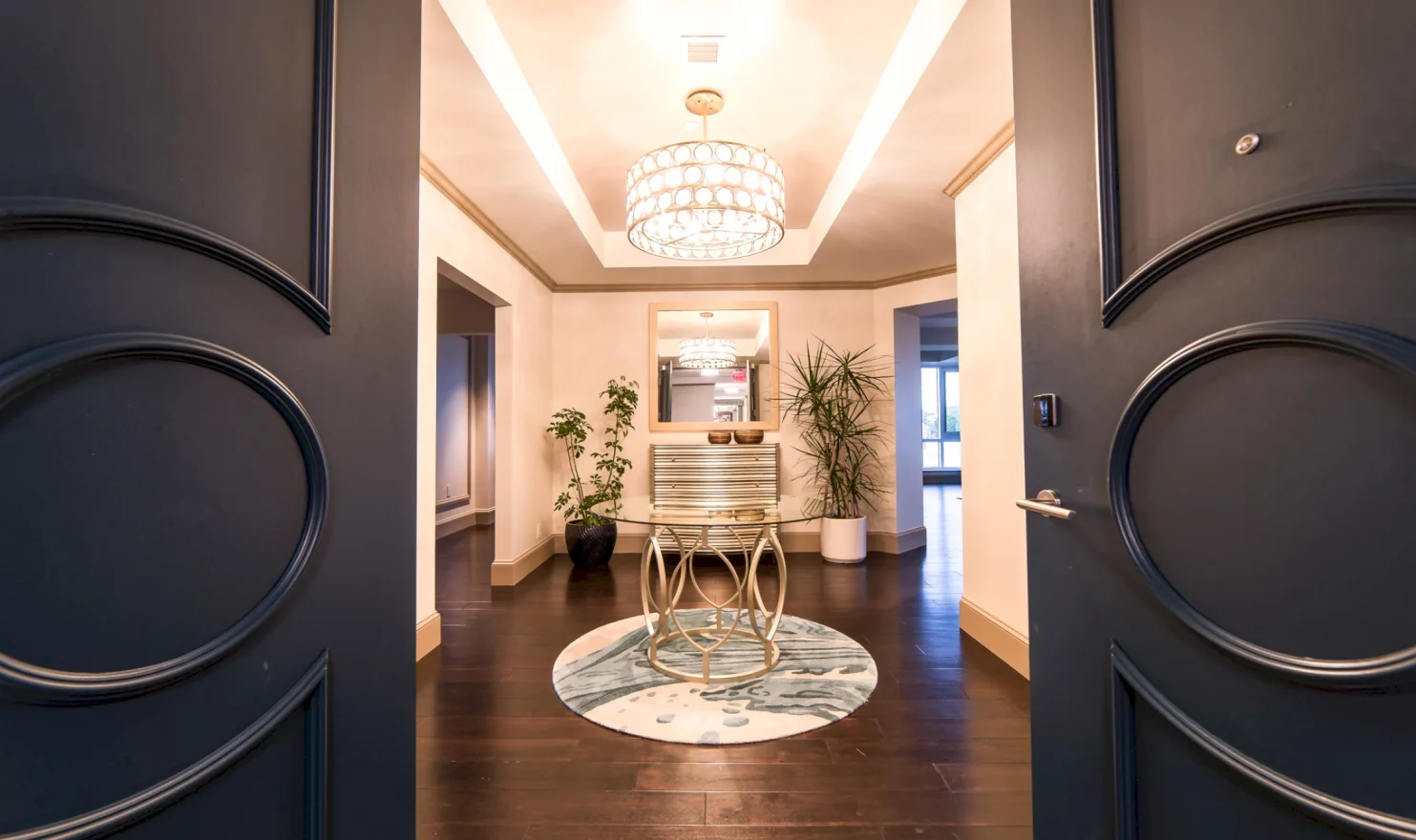 A round glass-top table and ornate chandelier are centered in a grand, brightly lit entryway, viewed through open dark doors, featuring dark wood floors and potted plants.