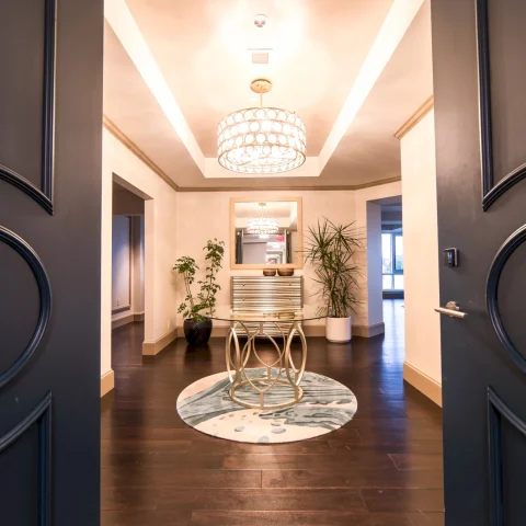 A round glass-top table and ornate chandelier are centered in a grand, brightly lit entryway, viewed through open dark doors, featuring dark wood floors and potted plants.
