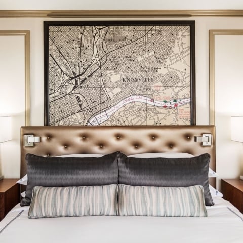 A bed is centered with a tufted headboard, dark and striped pillows. Above, a framed map depicts "KNOXVILLE", "UNIVERSITY OF TENNESSEE", "FORT SANDERS", "Adair Park", "Hampton Park". Modern lamps flank it on nightstands.