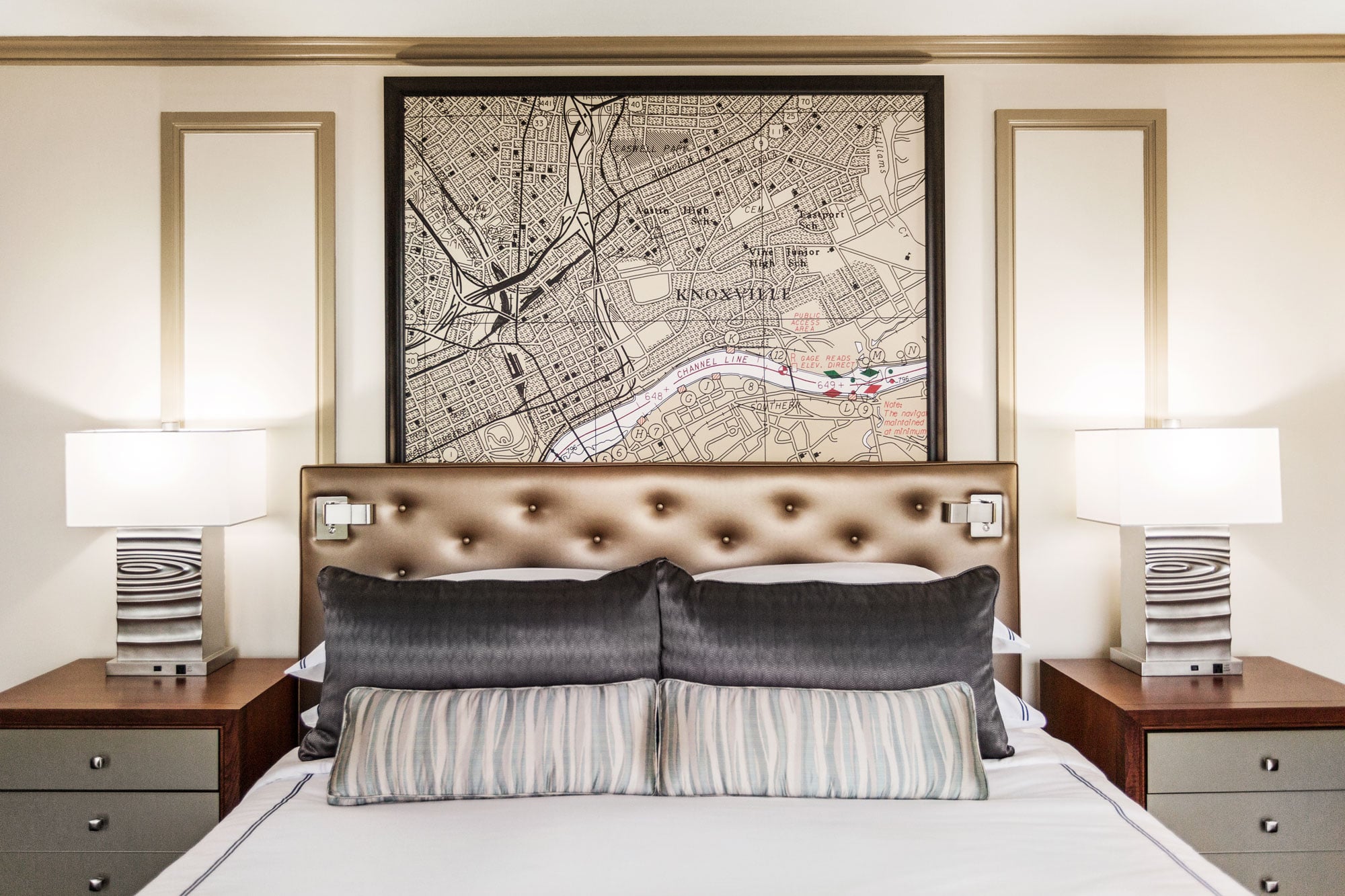 A bed is centered with a tufted headboard, dark and striped pillows. Above, a framed map depicts "KNOXVILLE", "UNIVERSITY OF TENNESSEE", "FORT SANDERS", "Adair Park", "Hampton Park". Modern lamps flank it on nightstands.