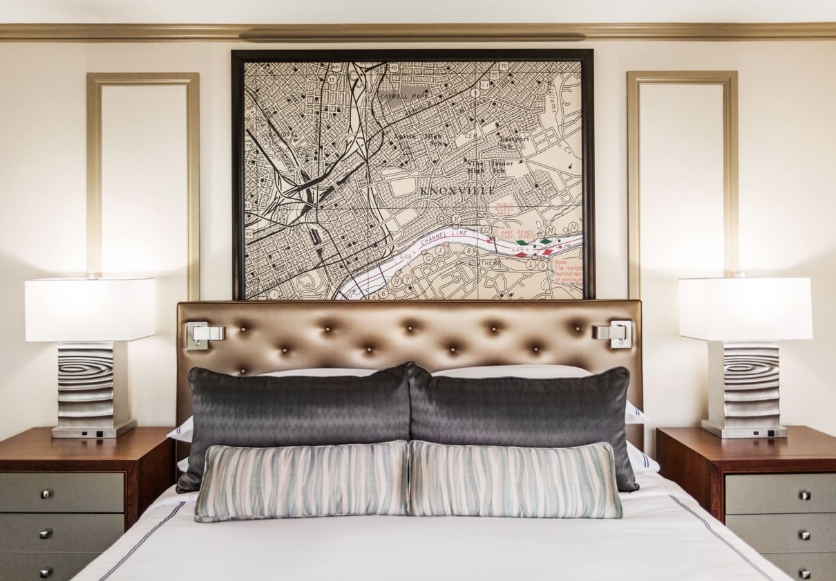 A neatly made bed with a brown tufted headboard is positioned under a large framed map of "KNOXVILLE". Two lamps sit on nightstands flanking the bed.