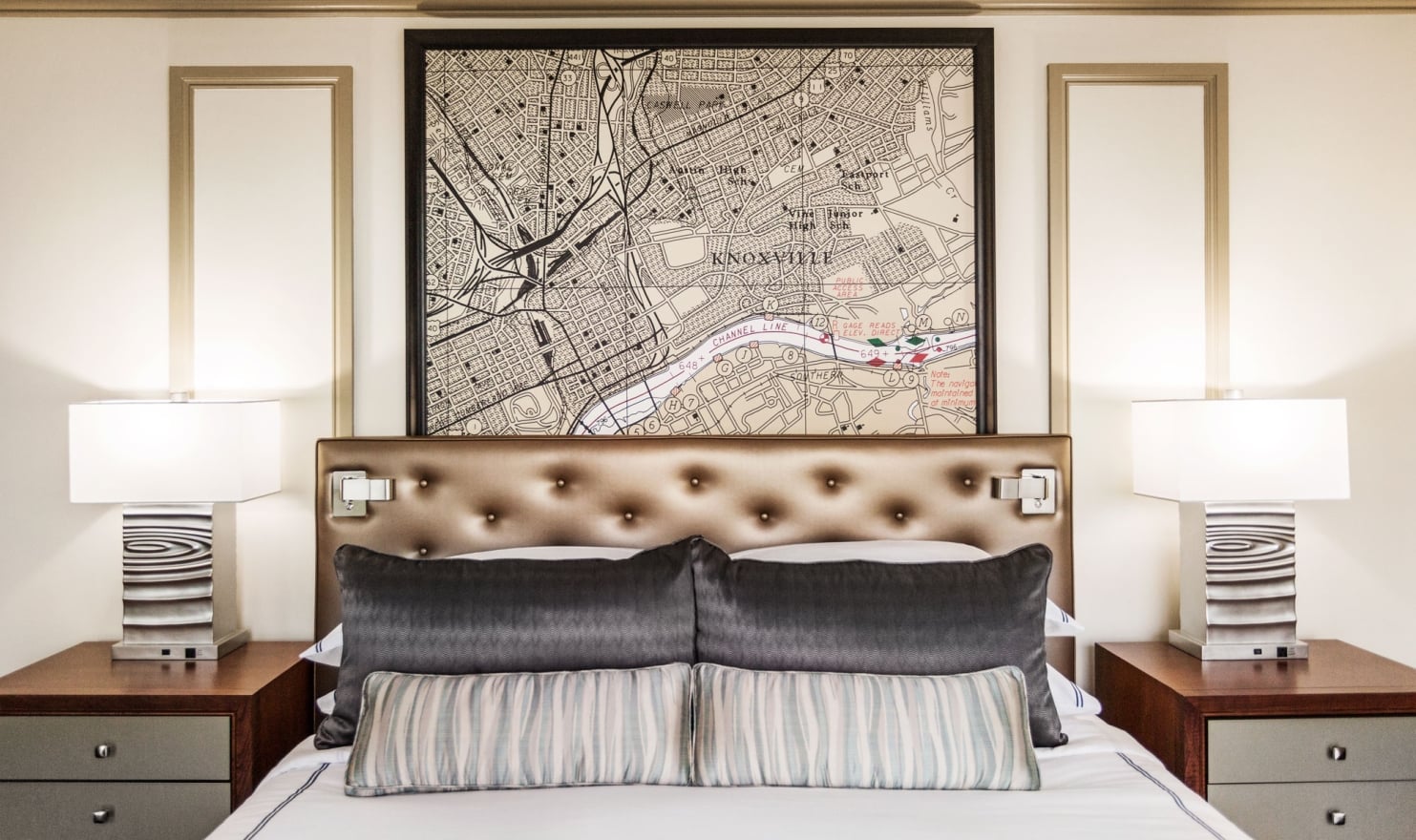 A neatly made bed with a brown tufted headboard is positioned under a large framed map of "KNOXVILLE". Two lamps sit on nightstands flanking the bed.