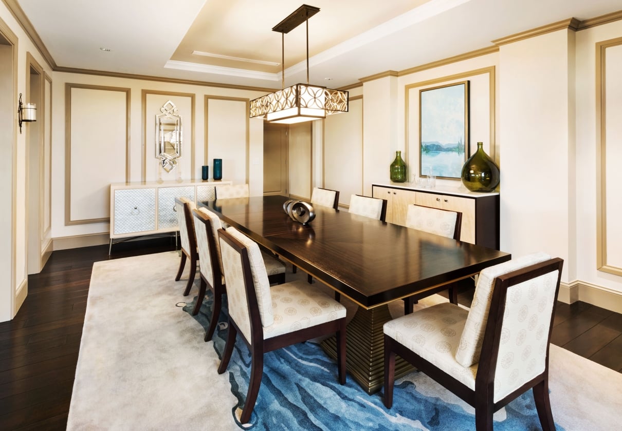 A long dark wood dining table with eight upholstered chairs sits on a blue-patterned rug in a luxurious room with ornate walls, sideboards, and a decorative chandelier.