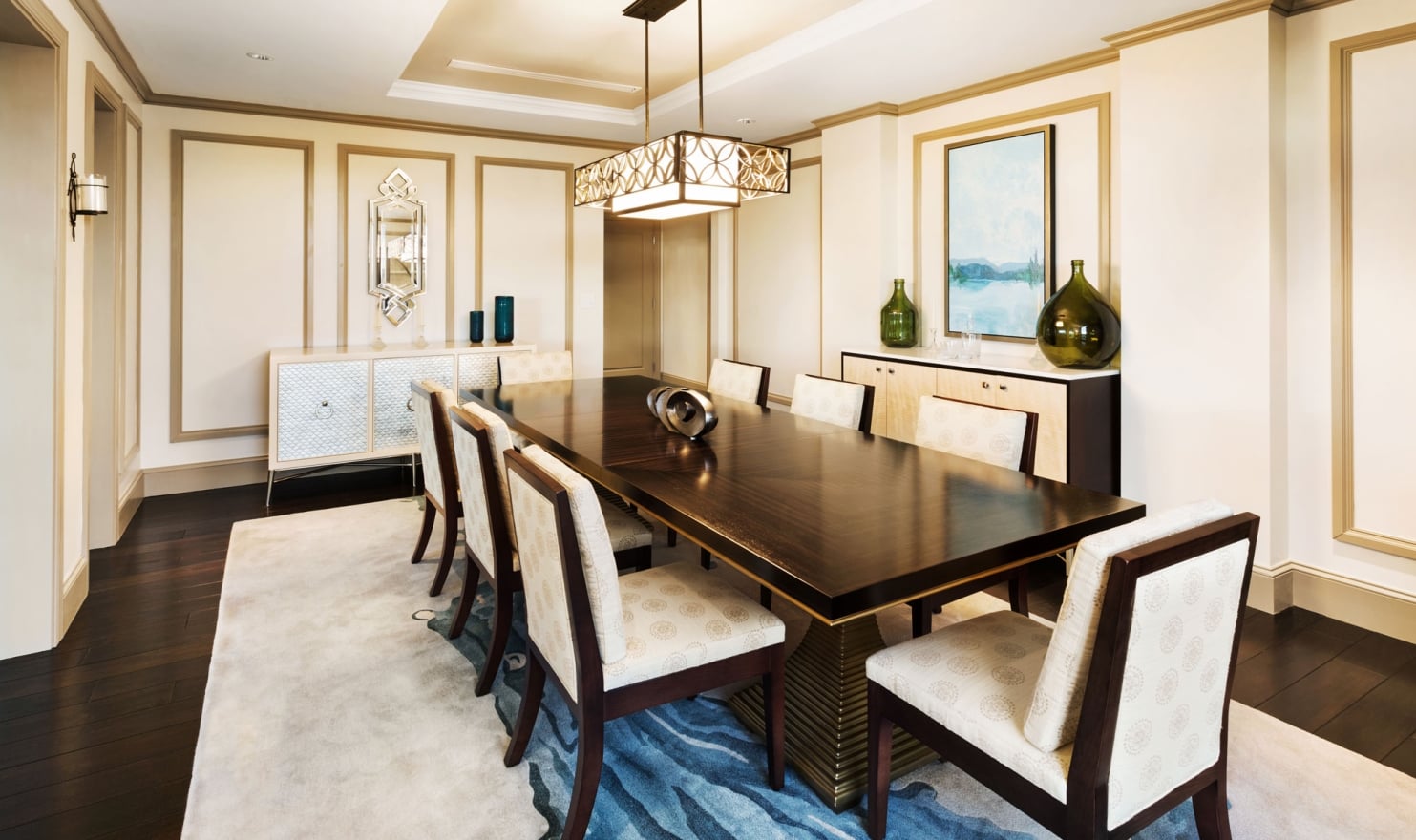 A long dark wood dining table with eight upholstered chairs sits on a blue-patterned rug in a luxurious room with ornate walls, sideboards, and a decorative chandelier.