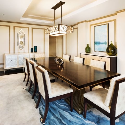 A long dark wood dining table with eight upholstered chairs sits on a blue-patterned rug in a luxurious room with ornate walls, sideboards, and a decorative chandelier.