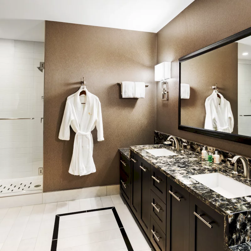 A white bathrobe hangs on a brown wall in a luxurious bathroom featuring dual vanities with dark granite counters, a large mirror, and a glass-enclosed shower.