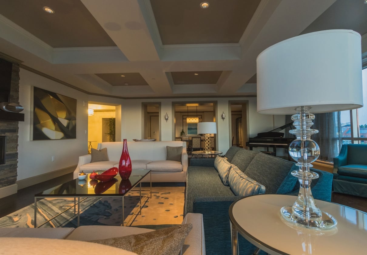 A luxurious living room features beige and teal sofas, a glass coffee table with red vases, and a Yamaha grand piano, in a spacious interior with decorative lighting and large windows.